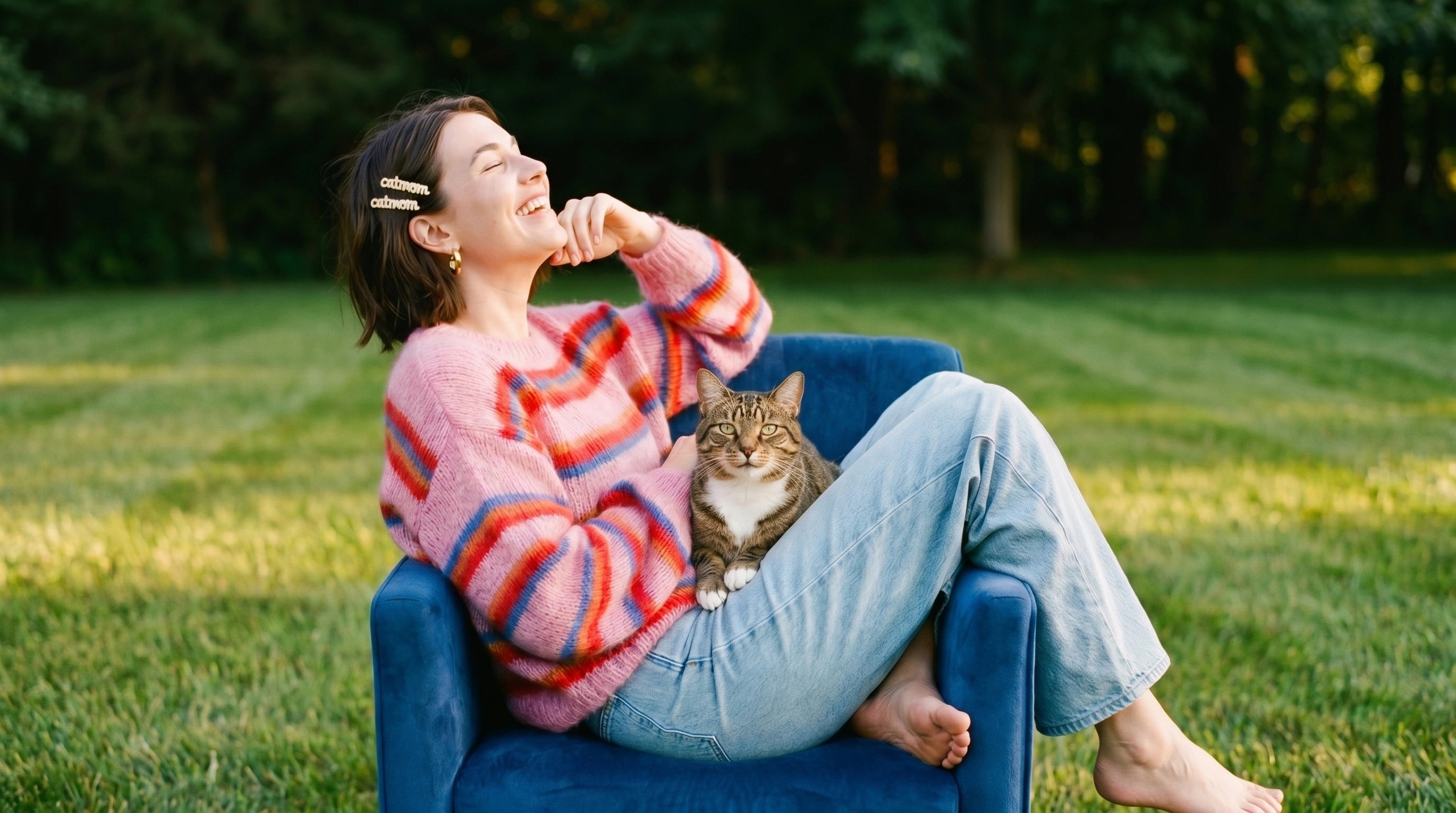 Woman sitting on a blue chair with a cat in her lap, surrounded by green grass and trees.